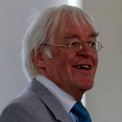 A portrait photograph of a man with white hair wearing a suit jackets, shirt and tie. He is wearing glasses and smiling