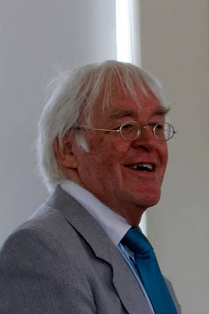 A portrait photograph of a man with white hair wearing a suit jackets, shirt and tie. He is wearing glasses and smiling