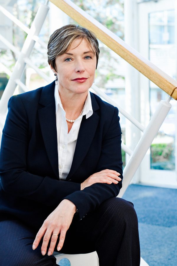 A woman in an office sat on some stairs. She is wearing a dark suita nd light blouse and has short highlighted hair.