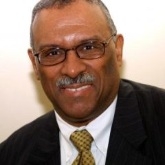 A man with glasses wearing a black suit jacket, white shirt and yellow tie