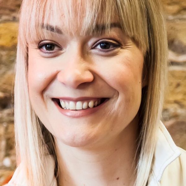 A woman with long fair hair and a fringe smiling. It is a portrait style photo. She has a brick wall behind her.