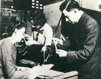 two women and one man working on an early computer. A man reads ticker tape amd a woman wearing glasses takes notes. Another women in the background is reading a report