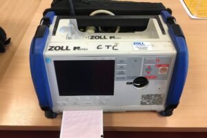 A colour photograph of a defibrillator with graph paper coming out of the front. It has a handle and a screen on the front and is a large grey rectangular box with blue sides