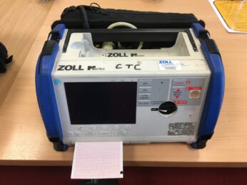 A colour photograph of a defibrillator with graph paper coming out of the front. It has a handle and a screen on the front and is a large grey rectangular box with blue sides
