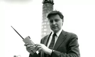 A man in a suit holding a vintage mobile telephone in front of the BT tower in the 1980s.