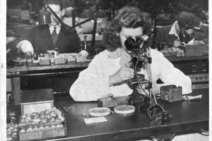 A woman sat at a bench with nails and other parts on the desk top, inspecting electrical equipment through a microscope.