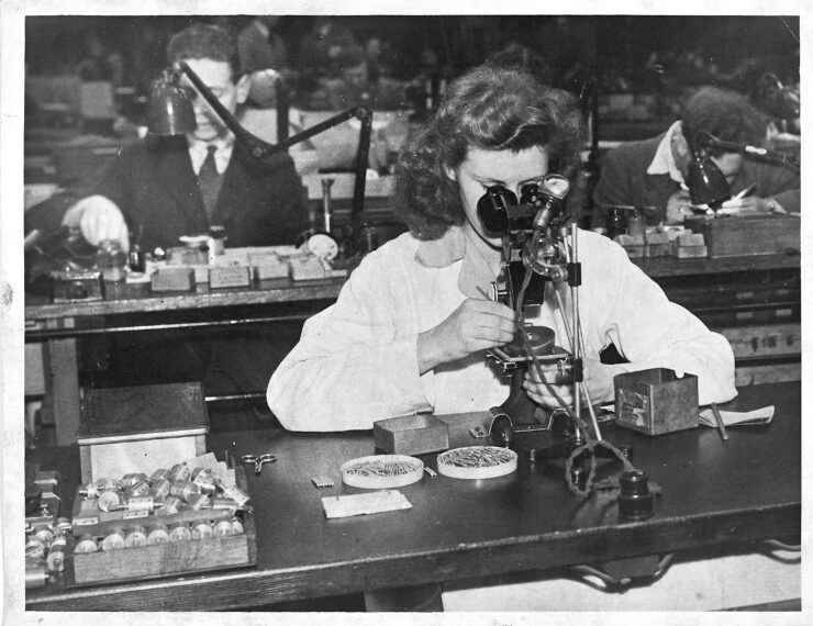 A woman sat at a bench with nails and other parts on the desk top, inspecting electrical equipment through a microscope.