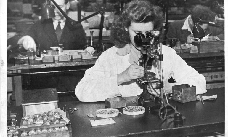 A woman sat at a bench with nails and other parts on the desk top, inspecting electrical equipment through a microscope.