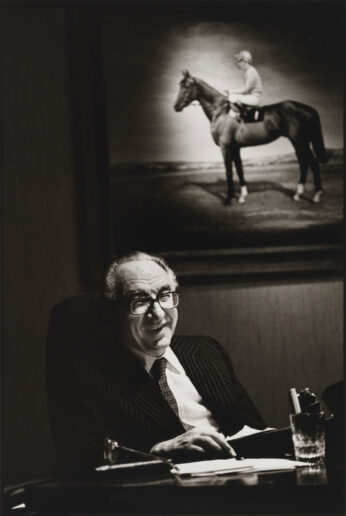 A man sat at a desk with a portrait of a horse on the wall behind him