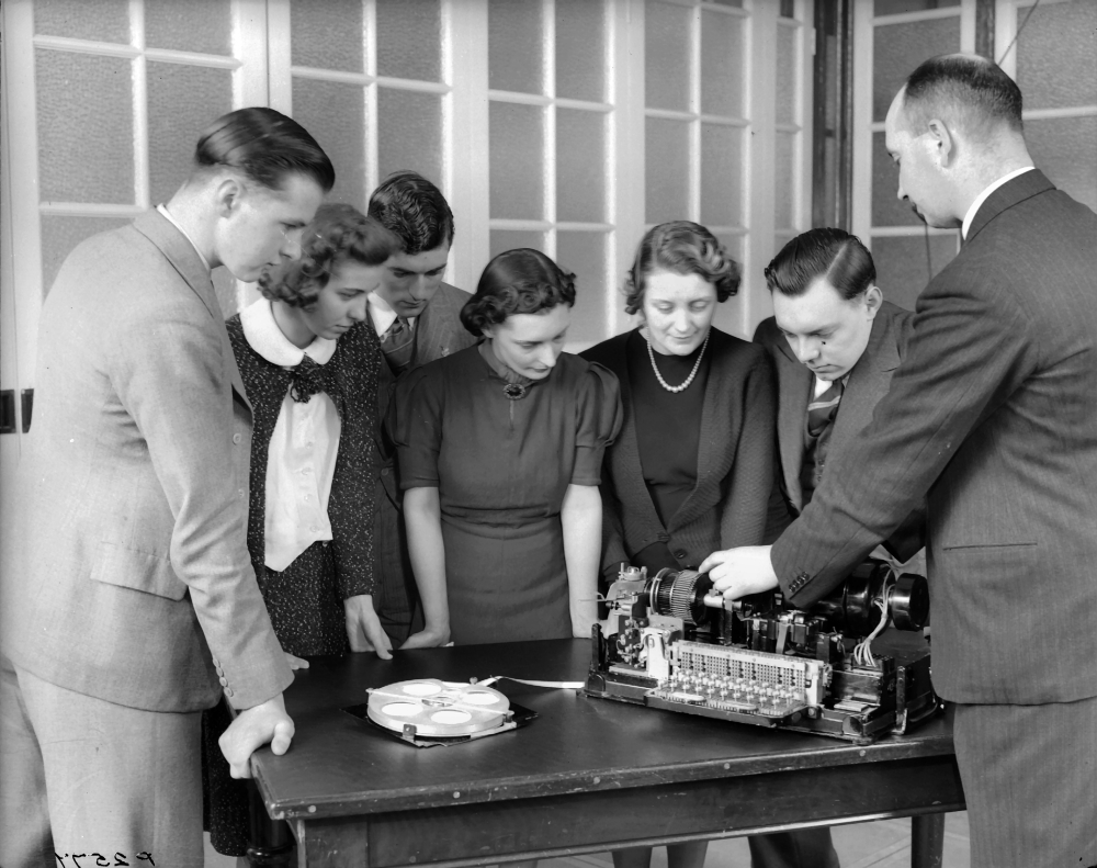 A group of young men and women in early twentieth century dress being instructed by a man in a dark suit about a machine on a desk.