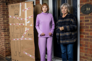 Humanoid robot Ann Droid stands next to Sue Johnston and a large cardboard box on a doorstep